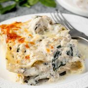A slice of creamy mushroom chicken Alfredo lasagna on a white plate with melted cheese and spinach, served with a fork.