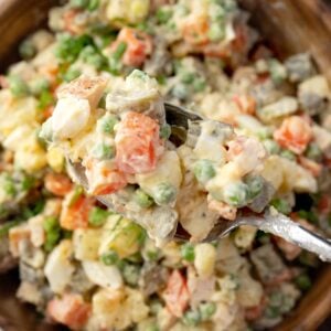 Close-up of Russian Salad Olivier on a spoon, showing creamy potato salad with carrots, peas, eggs, pickles, and turkey ham in a mayonnaise dressing.