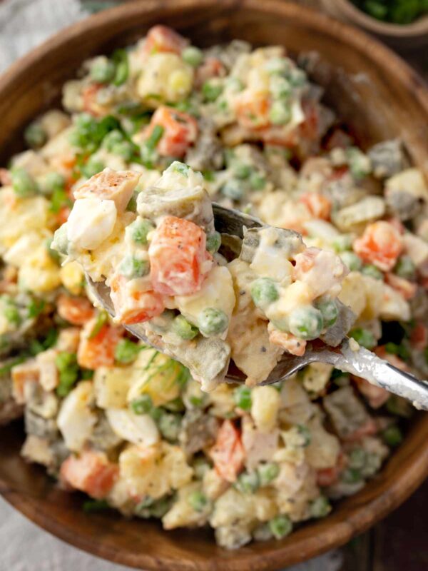 Close-up of Russian Salad Olivier on a spoon, showing creamy potato salad with carrots, peas, eggs, pickles, and turkey ham in a mayonnaise dressing.