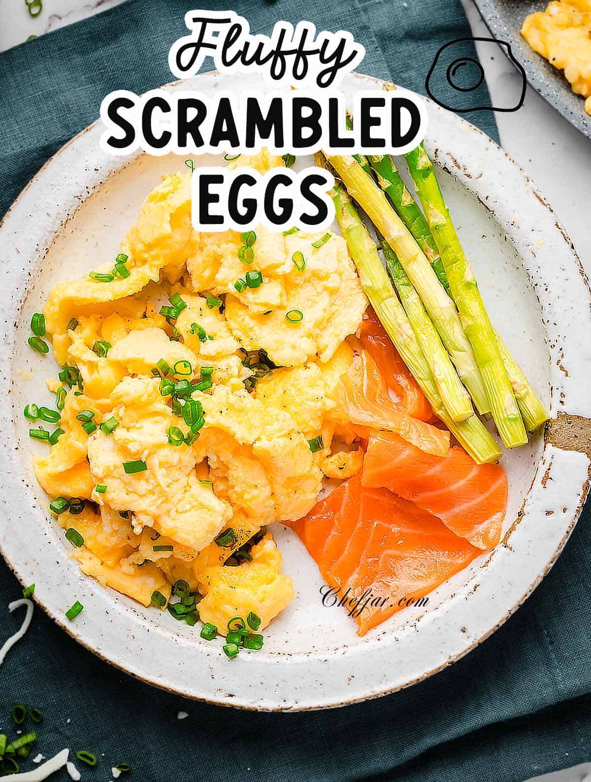A white platter with fluffy scrambled eggs with smoked salmon and roasted asparagus next to them. Garnished with green onions.