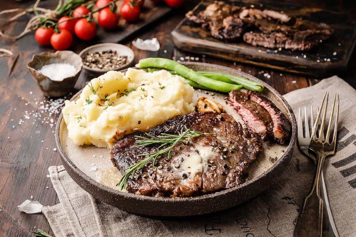 Pan seared wagyu ribeye steaks on a plate, served with mashed potatoes and roasted green beans. There are cherry tomatoes and a chopping board with a sliced wagyu steak in the background.