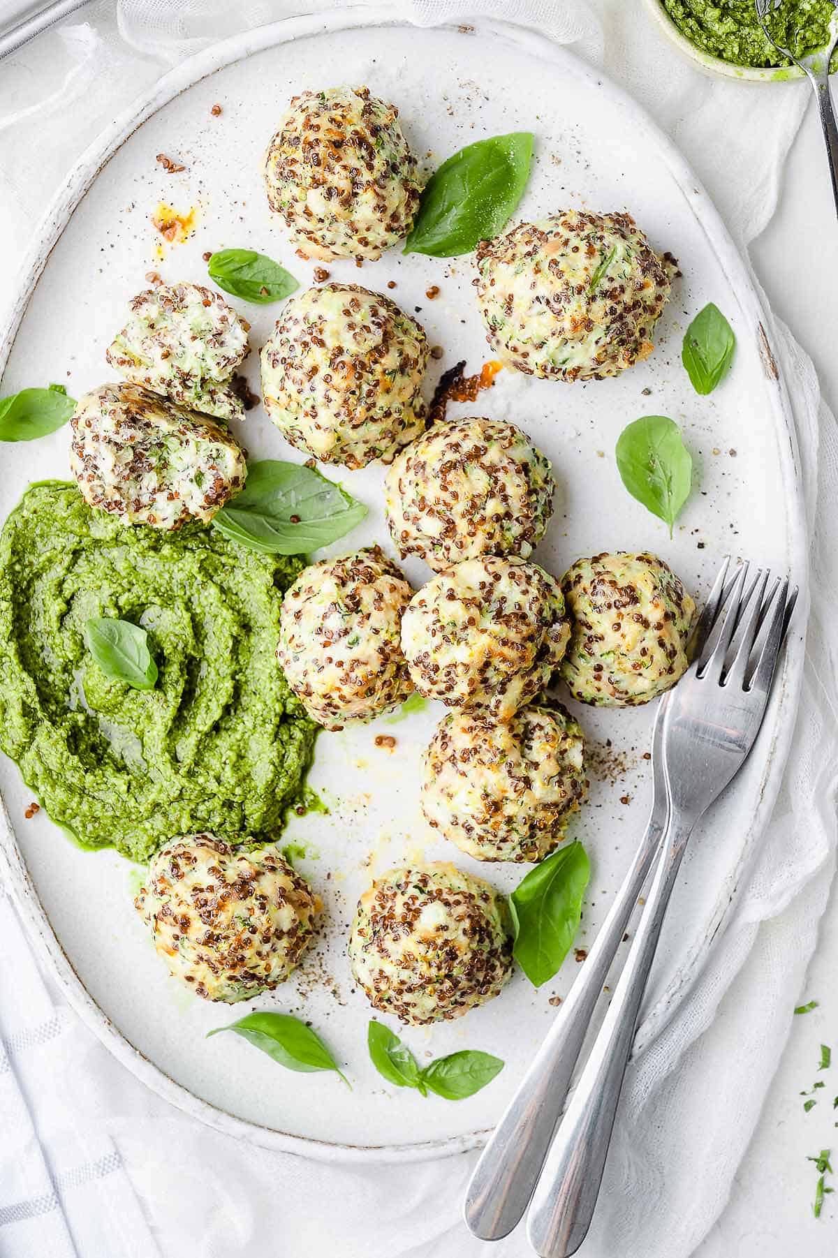 Chicken zucchini meatballs on a large oval white plate. Garnished with fresh basil leaves and served with pesto.