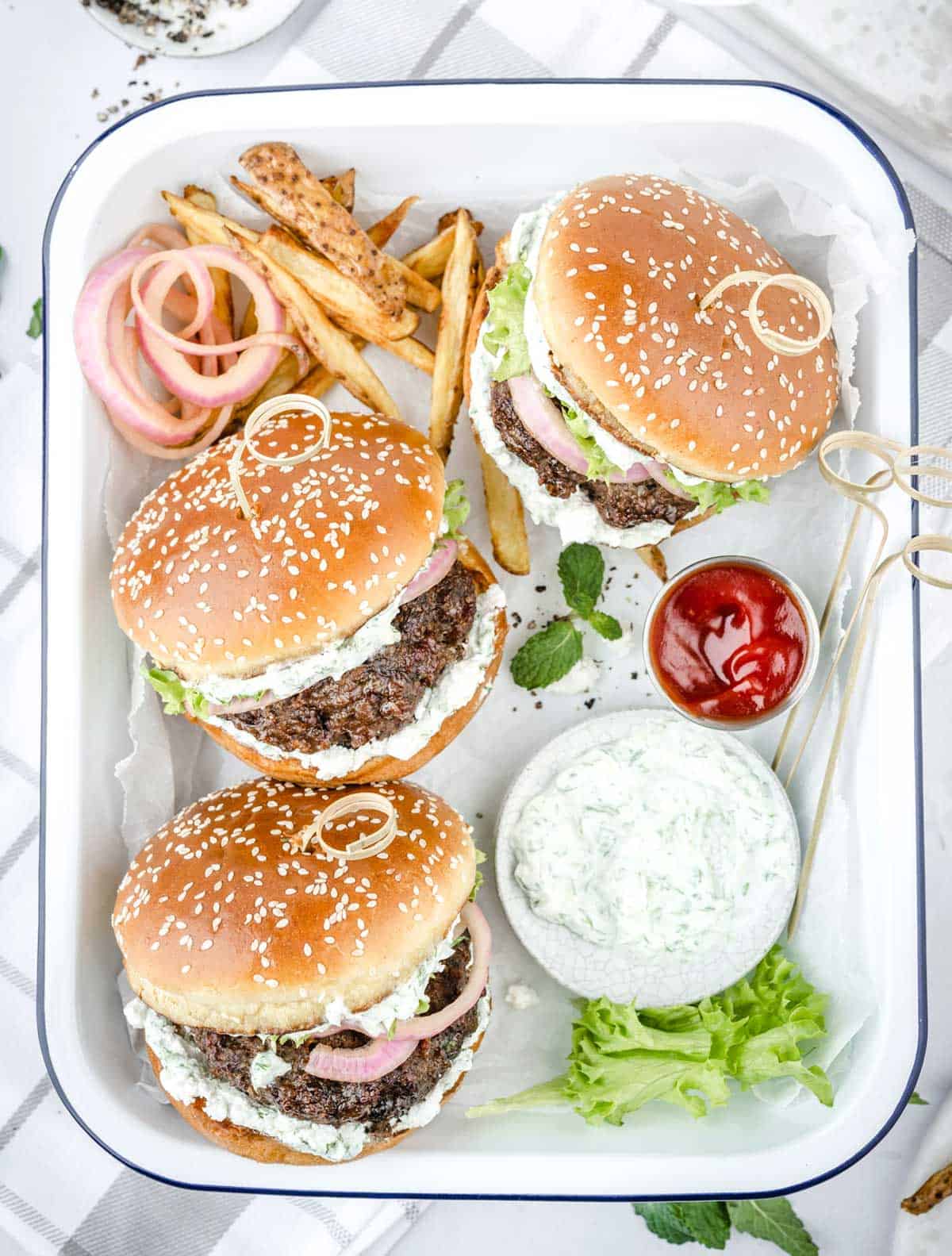 Three lamb burgers are served on a tray with French fries, tzatziki sauce and ketchup