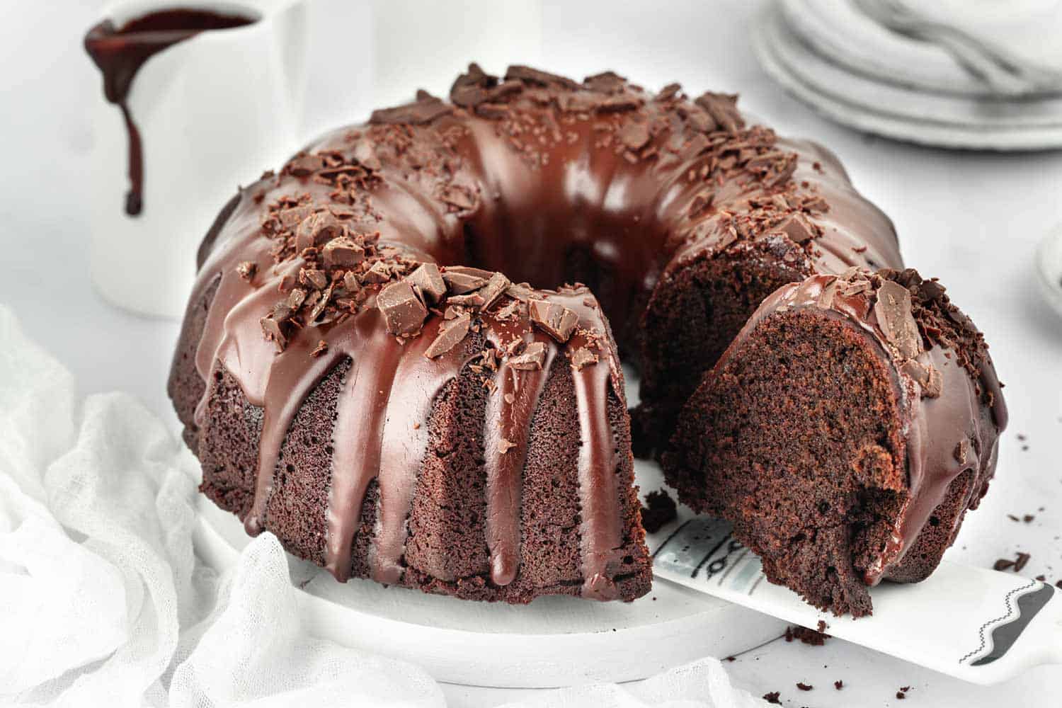 A slice of chocolate bundt cake being served from a cake stand with the whole cake on it.