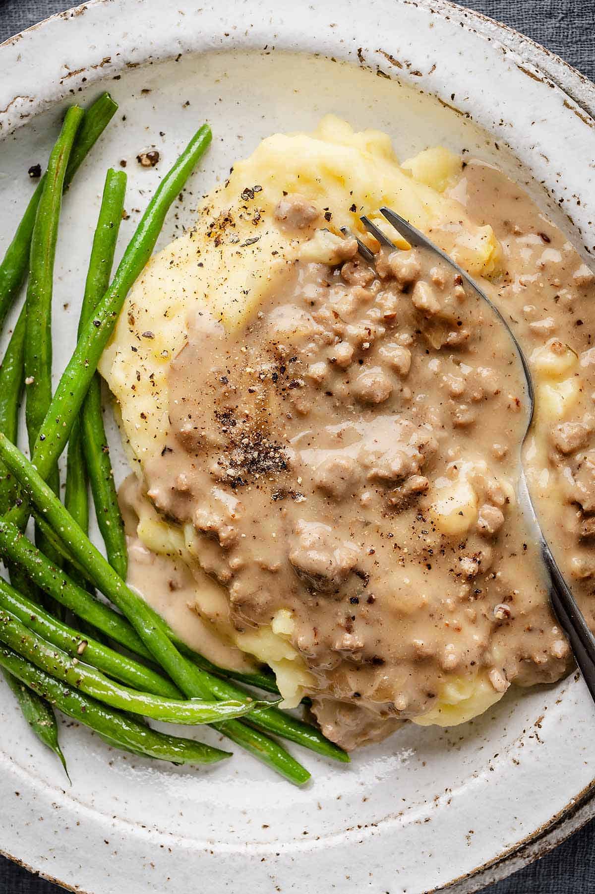 A pile of potatoes topped with Hamburger Gravy on a plate with green beans.