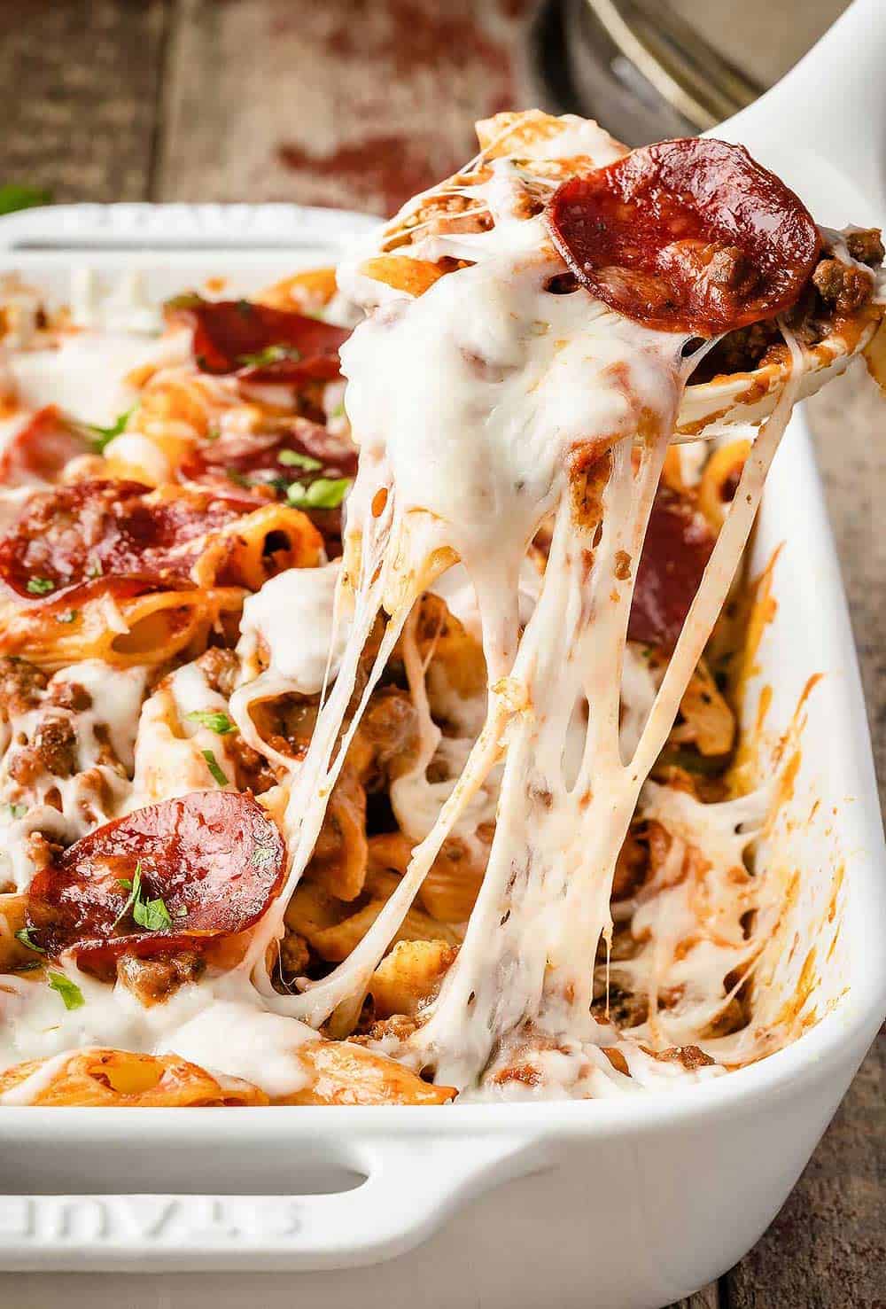 Pizza pasta in a white casserole dish with a spoon lifting up a serving out of the dish.
