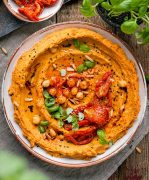 An overhead shot of white bowl of sun dried tomato hummus surrounded by sun dried tomatoes and basil / Sun Dried Tomato Hummus