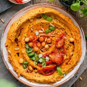 An overhead shot of white bowl of sun dried tomato hummus surrounded by sun dried tomatoes and basil / Sun Dried Tomato Hummus