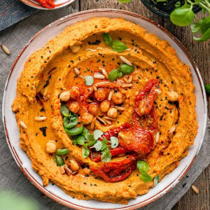 An overhead shot of white bowl of sun dried tomato hummus surrounded by sun dried tomatoes and basil / Sun Dried Tomato Hummus