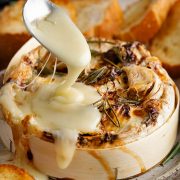 A spoon scooping melted Camembert cheese from a baked wheel, with strings of cheese dripping down.