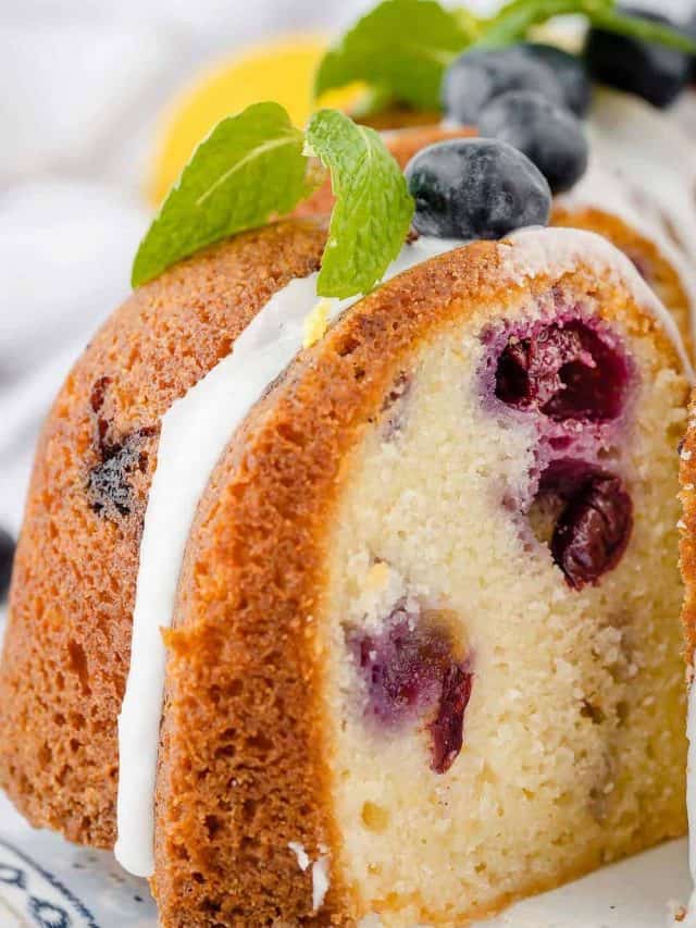 Removing a slice of lemon blueberry bundt cake from the whole cake. The cake is golden brown and looks moist and tender with blueberries visible throughout.