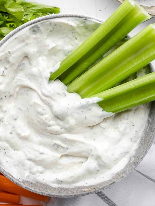 Texas Roadhouse Ranch dressing in a grey bowl with celery sticks on the side. The dressing is creamy and tangy, with specks of green herbs and spices visible. The celery sticks provide a colorful and crunchy contrast to the dressing.