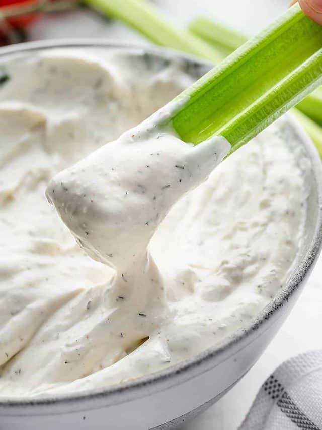 A hand holding a celery stick and dipping it into a bowl of Texas Roadhouse Ranch dressing. The dressing is creamy and tangy.