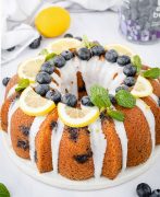 A photo of a lemon blueberry bundt cake on a white serving board. The cake is moist and golden brown, with small bursts of blueberries dotted throughout. On top of the cake, there are fresh blueberries, thin slices of lemon, and a sprig of mint leaves arranged in a decorative manner.