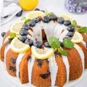 A photo of a lemon blueberry bundt cake on a white serving board. The cake is moist and golden brown, with small bursts of blueberries dotted throughout. On top of the cake, there are fresh blueberries, thin slices of lemon, and a sprig of mint leaves arranged in a decorative manner.