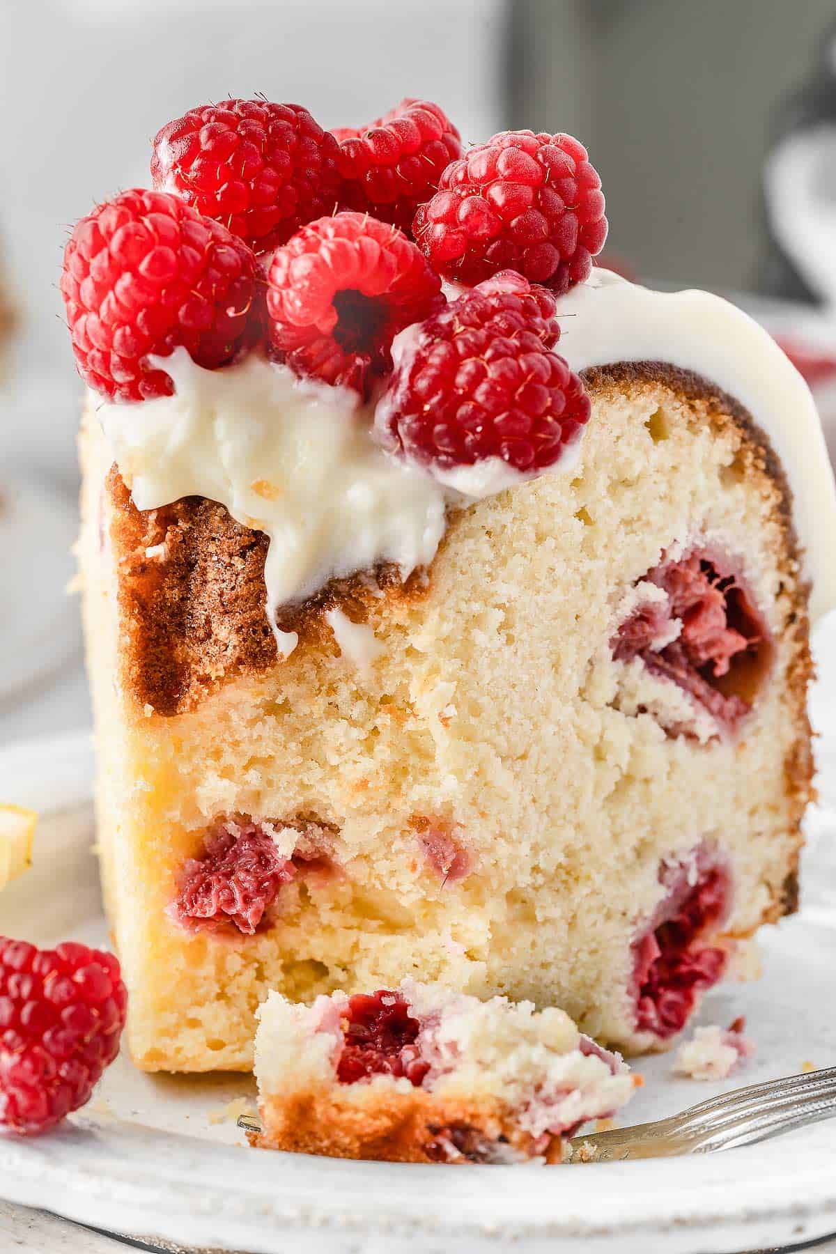Partially eaten slice of Lemon Raspberry Bundt Cake on a white plate with fresh raspberries and lemon slices on top.