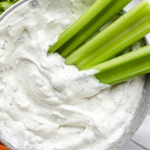 Texas Roadhouse Ranch dressing in a grey bowl with celery sticks on the side. The dressing is creamy and tangy, with specks of green herbs and spices visible. The celery sticks provide a colorful and crunchy contrast to the dressing.