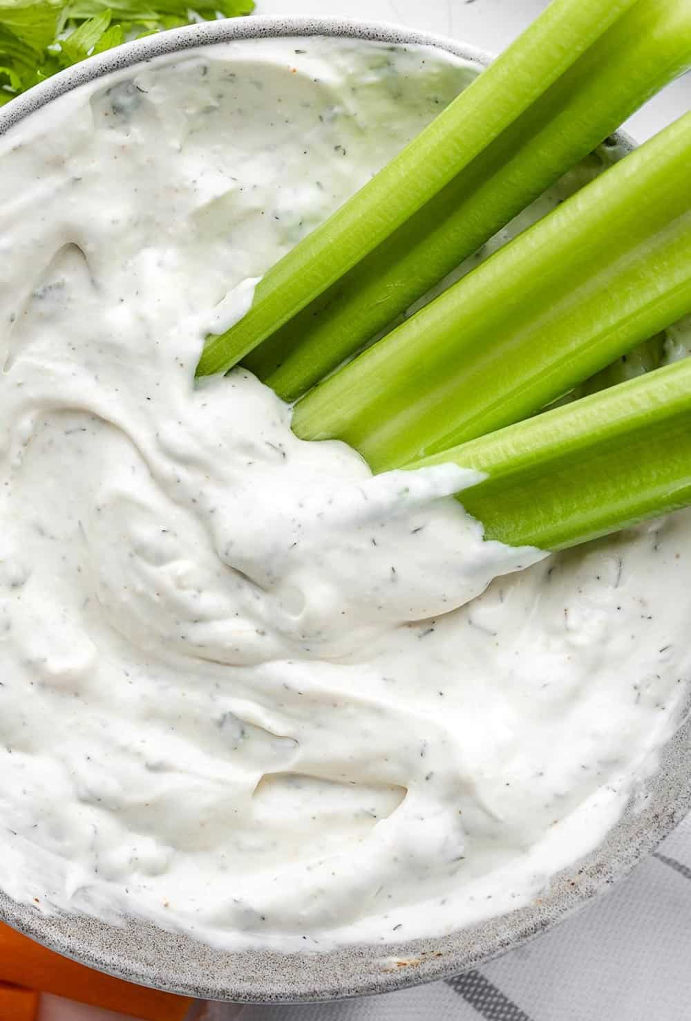 Texas Roadhouse Ranch dressing in a grey bowl with celery sticks on the side. The dressing is creamy and tangy, with specks of green herbs and spices visible. The celery sticks provide a colorful and crunchy contrast to the dressing.