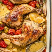 A golden-brown roasted chicken with crispy skin in a large roasting pan surrounded by cherry tomatoes, green olives, shallots, and garlic cloves. The pan juices are visible and a spoon is nearby for spooning the sauce over it.