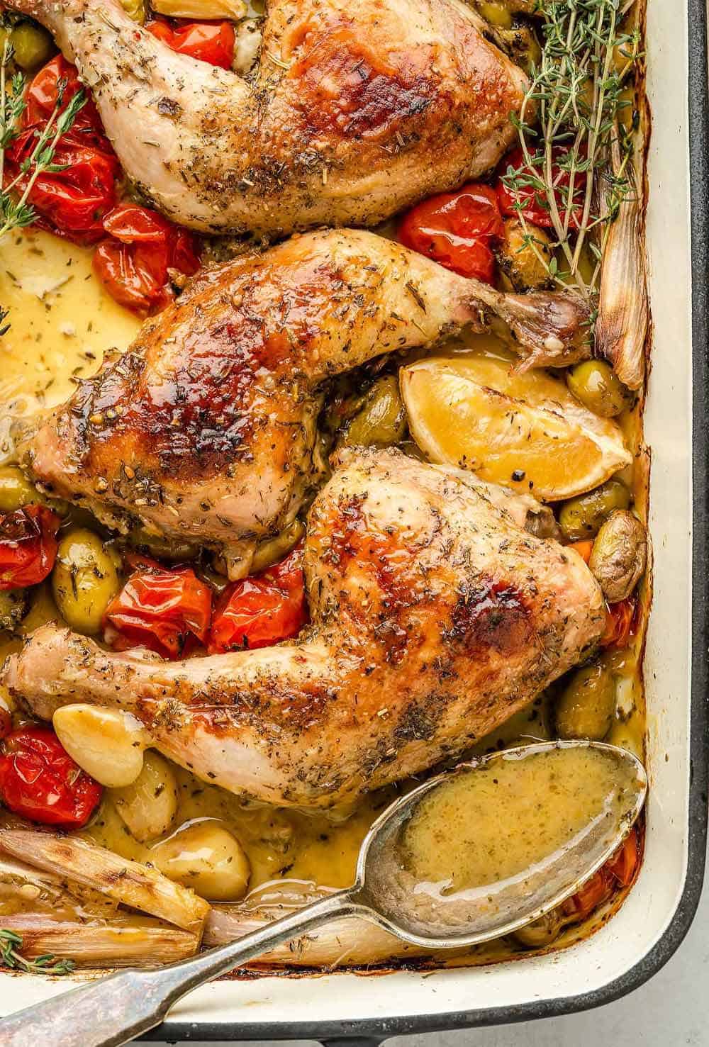 A golden-brown roasted chicken with crispy skin in a large roasting pan surrounded by cherry tomatoes, green olives, shallots, and garlic cloves. The pan juices are visible and a spoon is nearby for spooning the sauce over it.