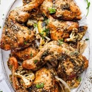 Za'atar chicken with roasted cauliflower, shallots, and lemon on an oval white plate. The chicken is golden brown with crispy skin and is served on a bed of roasted vegetables. The dish is garnished with fresh herbs and a drizzle of zaatar oil.