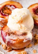 A close-up image of a peach half served on a white plate, topped with creamy vanilla ice cream and a generous sprinkling of crunchy granola.