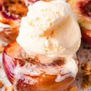 A close-up image of a peach half served on a white plate, topped with creamy vanilla ice cream and a generous sprinkling of crunchy granola.