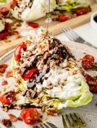Wedge salad on a plate being drizzled with balsamic vinegar glaze, topped with homemade blue dressing, tomatoes, onion, crispy bacon, and toasted chopped pecans.