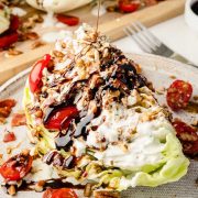 Wedge salad on a plate being drizzled with balsamic vinegar glaze, topped with homemade blue dressing, tomatoes, onion, crispy bacon, and toasted chopped pecans.