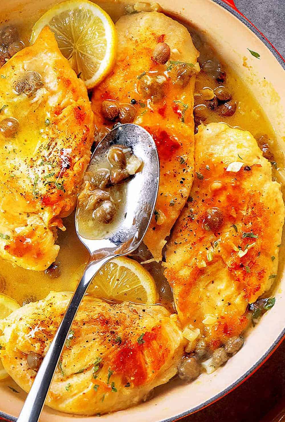 Image of chicken piccata being cooked in a skillet. The chicken is golden brown and has a crispy coating. The skillet contains a lemon sauce with capers and parsley, and there are slices of lemon and parsley leaves scattered around the chicken.