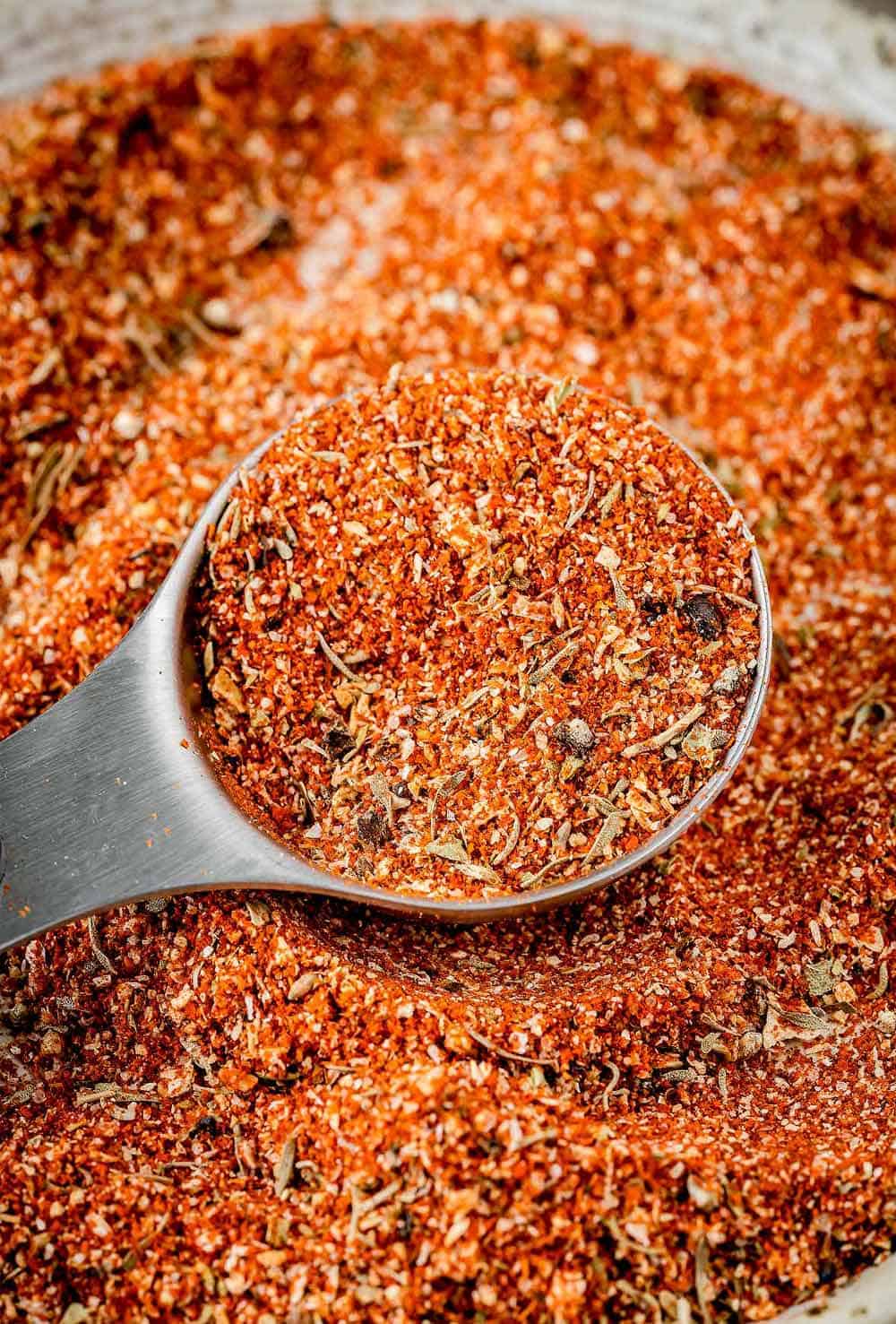 An image of homemade blackening seasoning on a white plate with a measuring spoon next to it. The seasoning is a dark reddish-brown color and is a mixture of various spices, including paprika, cayenne pepper, garlic powder, onion powder, thyme, and oregano. The spoon is filled with the seasoning.