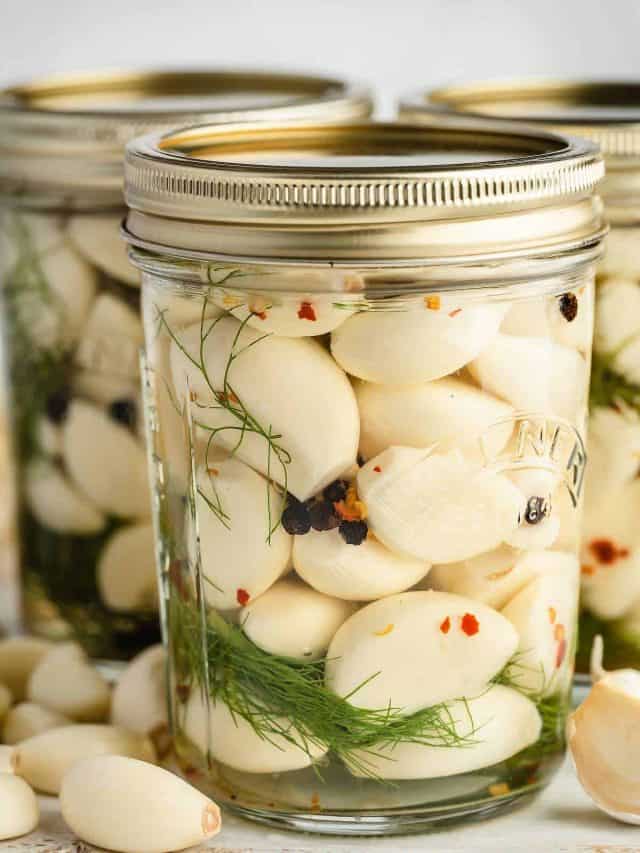 Close-up image of a wide mouth Kilner jar filled with pickled garlic cloves. The garlic cloves are submerged in a light-colored vinegar brine and are visible through the clear glass jar. In the background, two other jars can be seen.