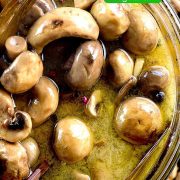 Overhead image of a glass bowl filled with marinated mushrooms in marinade. The mushrooms are brown and glossy, and the marinade contains visible herbs and spices.