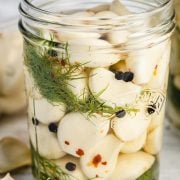 Image of a wide mouth Kilner jar filled with pickled garlic cloves. The garlic cloves are submerged in a light-colored vinegar brine and are visible through the clear glass jar.