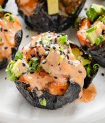 Close-up photo of a spicy salmon rice muffin topped with small pieces of avocado, sesame seeds, and a drizzle of spicy mayo.