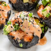 Close-up photo of a spicy salmon rice muffin topped with small pieces of avocado, sesame seeds, and a drizzle of spicy mayo.