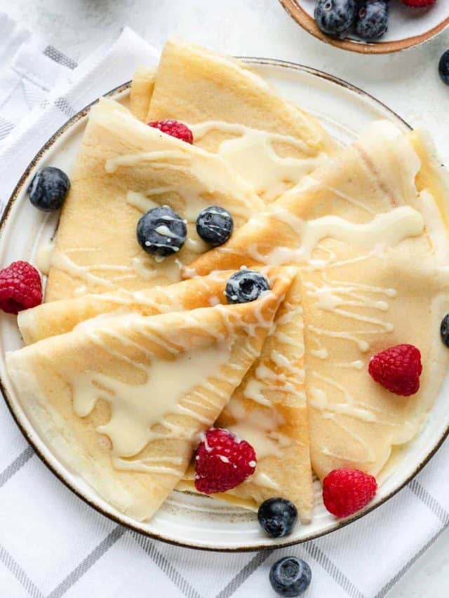 Easy homemade crepes on a white plate drizzled with condensed milk and topped with fresh berries.