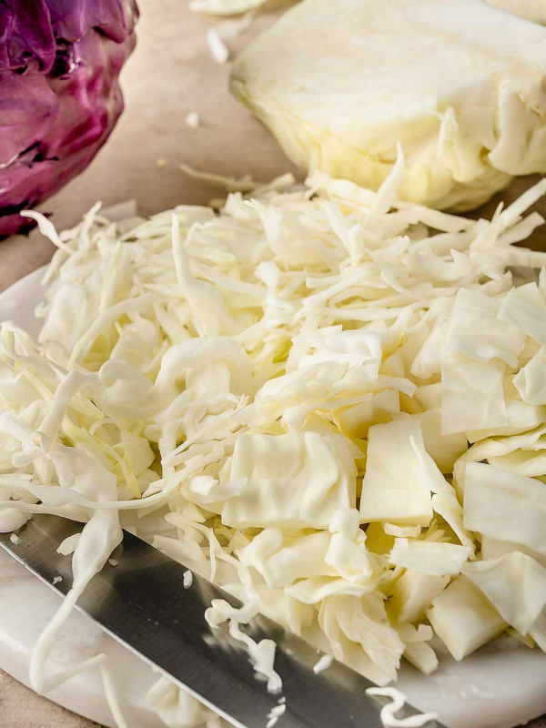 Image of freshly cut cabbage on a white cutting board, shredded and cut into thin squares, and cut into wedges, with a kitchen knife beside it.
