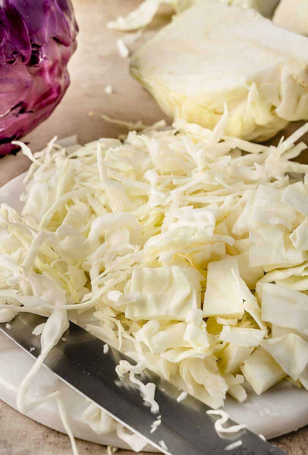 Image of freshly cut cabbage on a white cutting board, shredded and cut into thin squares, and cut into wedges, with a kitchen knife beside it.