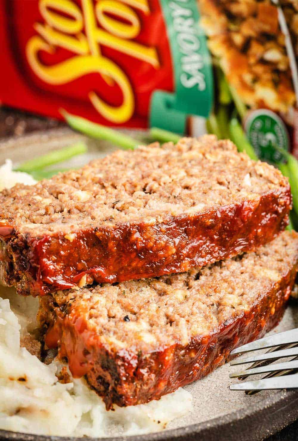 The photo shows two slices of meatloaf made with Stove Top stuffing mix, served on a plate alongside mashed potatoes and green beans. In the background, there is a box of Stove Top stuffing mix. The meatloaf appears moist and flavorful with a golden-brown crust.