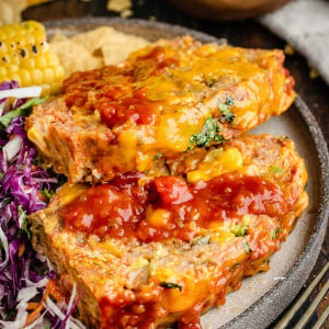Two slices of Taco meatloaf topped with taco sauce, accompanied by cilantro lime slaw and corn on the cob.