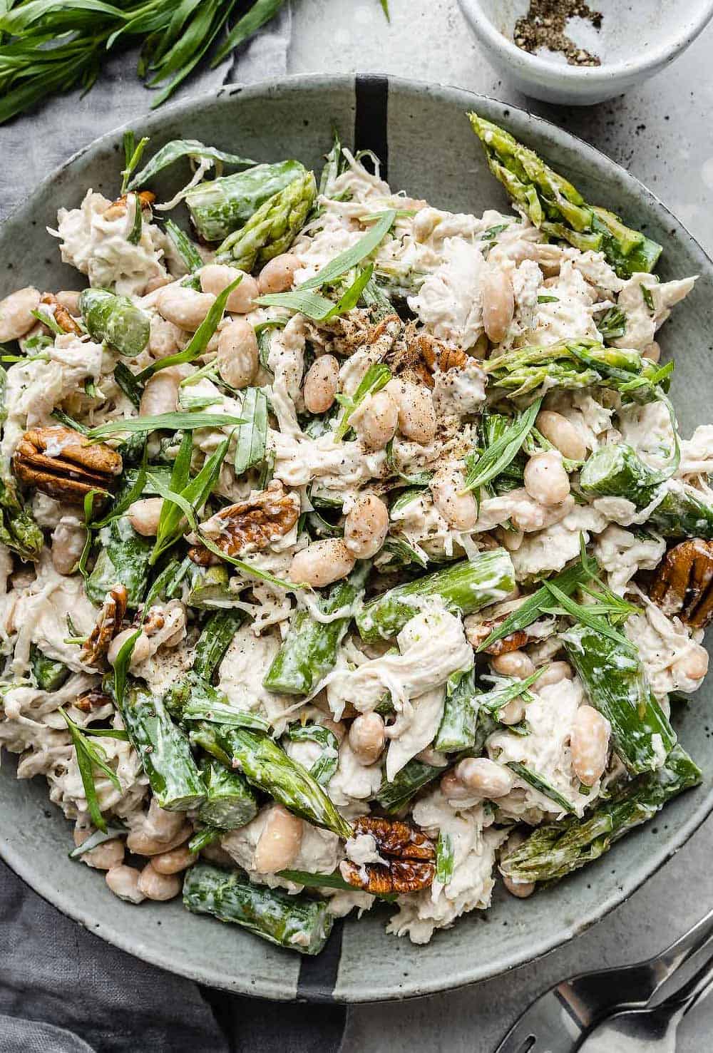 Tarragon Chicken Salad in a grey bowl: Shredded chicken mixed with asparagus, beans, nuts, and tarragon leaves, coated in a creamy dressing.