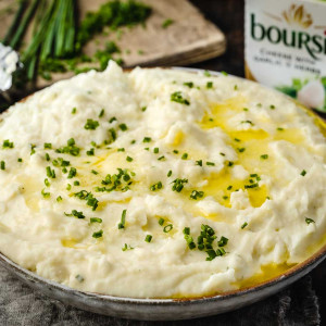 Boursin Mashed Potatoes in a bowl . Chopped chives and Boursin cheese in the background.