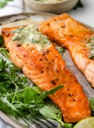 Two perfectly pan-seared salmon fillets with a golden-brown crust, served on a white plate. The fillets are accompanied by mixed salad leaves.