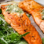 Two perfectly pan-seared salmon fillets with a golden-brown crust, served on a white plate. The fillets are accompanied by mixed salad leaves.