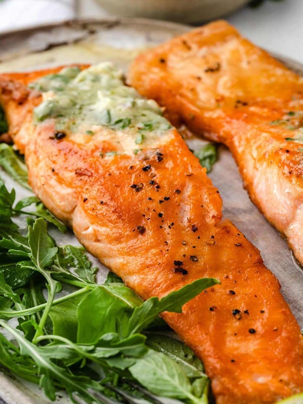 Two perfectly pan-seared salmon fillets with a golden-brown crust, served on a white plate. The fillets are accompanied by mixed salad leaves.