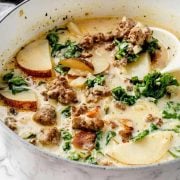 Image of Zuppa Toscana Soup in a Dutch oven. The soup is creamy and abundant with Italian sausage, potato slices, and kale, showcasing a variety of colors and textures.