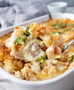 A hand holding a serving spoon lifting a portion of chicken cordon bleu casserole, revealing layers of chicken, ham, and melted cheese topped with a golden-brown breadcrumb crust.