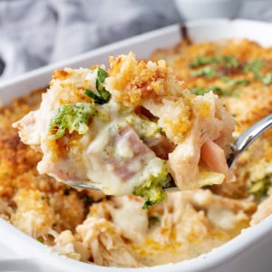 A hand holding a serving spoon lifting a portion of chicken cordon bleu casserole, revealing layers of chicken, ham, and melted cheese topped with a golden-brown breadcrumb crust.