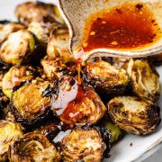 Pouring sauce over a dish of crispy, golden-brown Brussels sprouts. The sauce is drizzling evenly, adding a glossy sheen to the roasted sprouts.